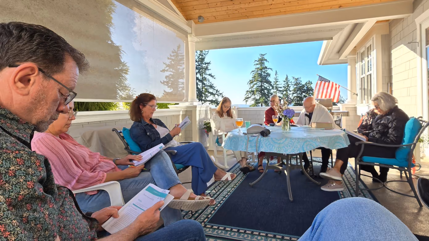 Group of seven adults seated on a covered patio in a circle, reading papers or booklets, with a table in the center holding drinks and flowers, an American flag visible outside.