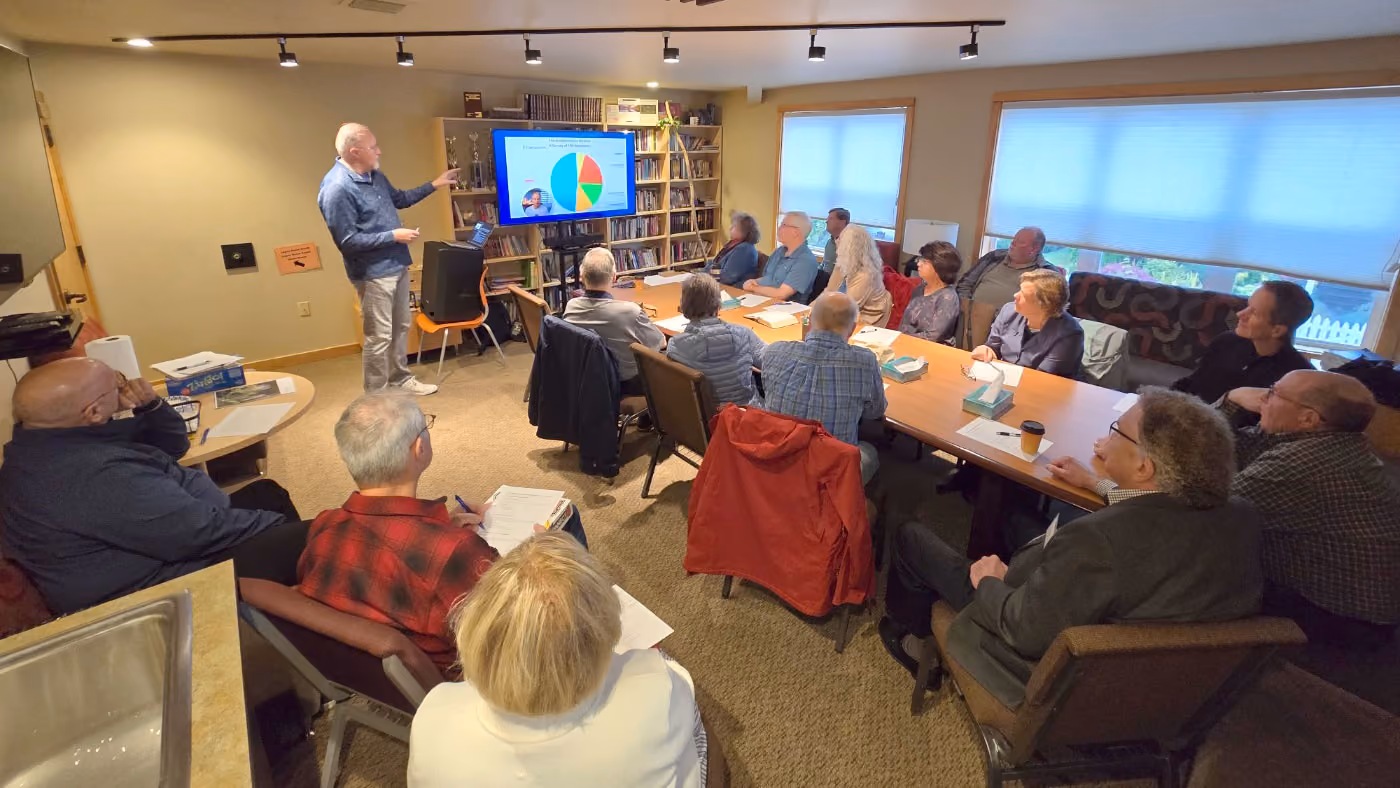 A man standing and pointing at a screen with a pie chart in a room where about 15 adults are seated around tables, attentively watching the presentation.