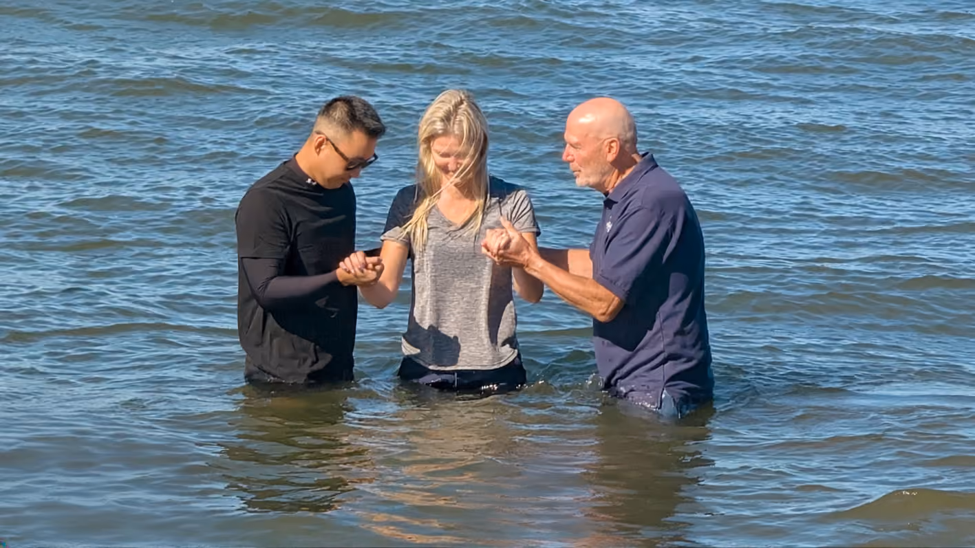 Three people standing waist-deep in water during a baptism ceremony, with a young woman in the center holding hands with two men on either side.