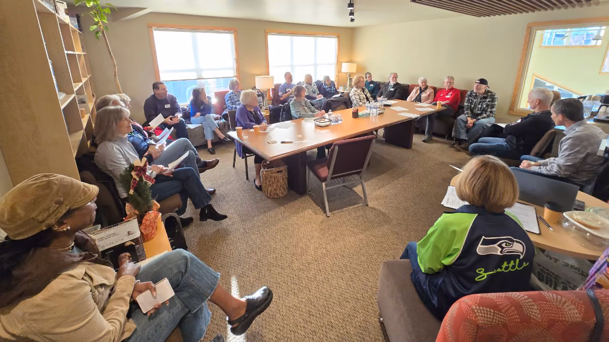 A diverse group of around twenty people seated in a large rectangular room, arranged around and beside a large wooden table, engaged in a meeting or discussion, with sunlight coming through two large windows.