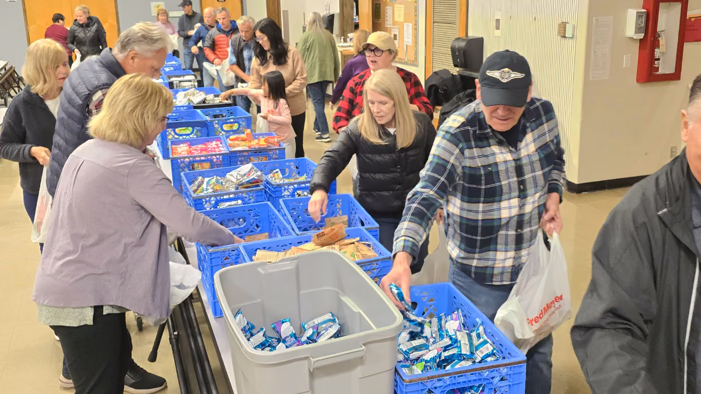 A group of people standing in line indoors, picking packaged food items from blue crates placed on tables, participating in a community food distribution event.