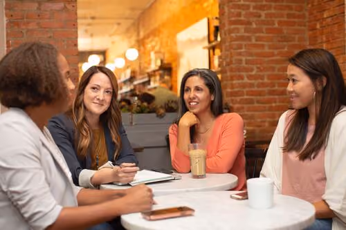 Four women sitting around two white tables in a cozy cafe with exposed brick walls, engaged in conversation with drinks on the table.