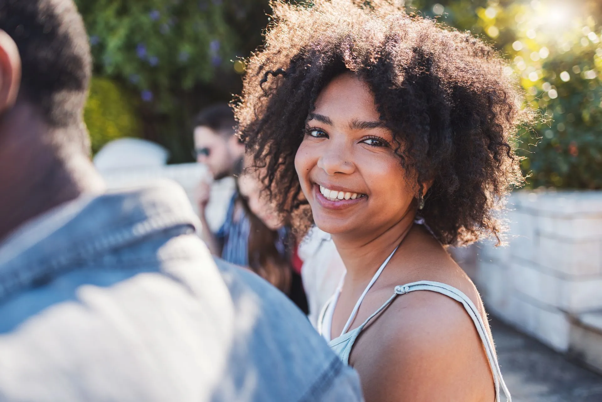 Smiling woman with curly hair outdoors in sunlight, with blurred people in the background.