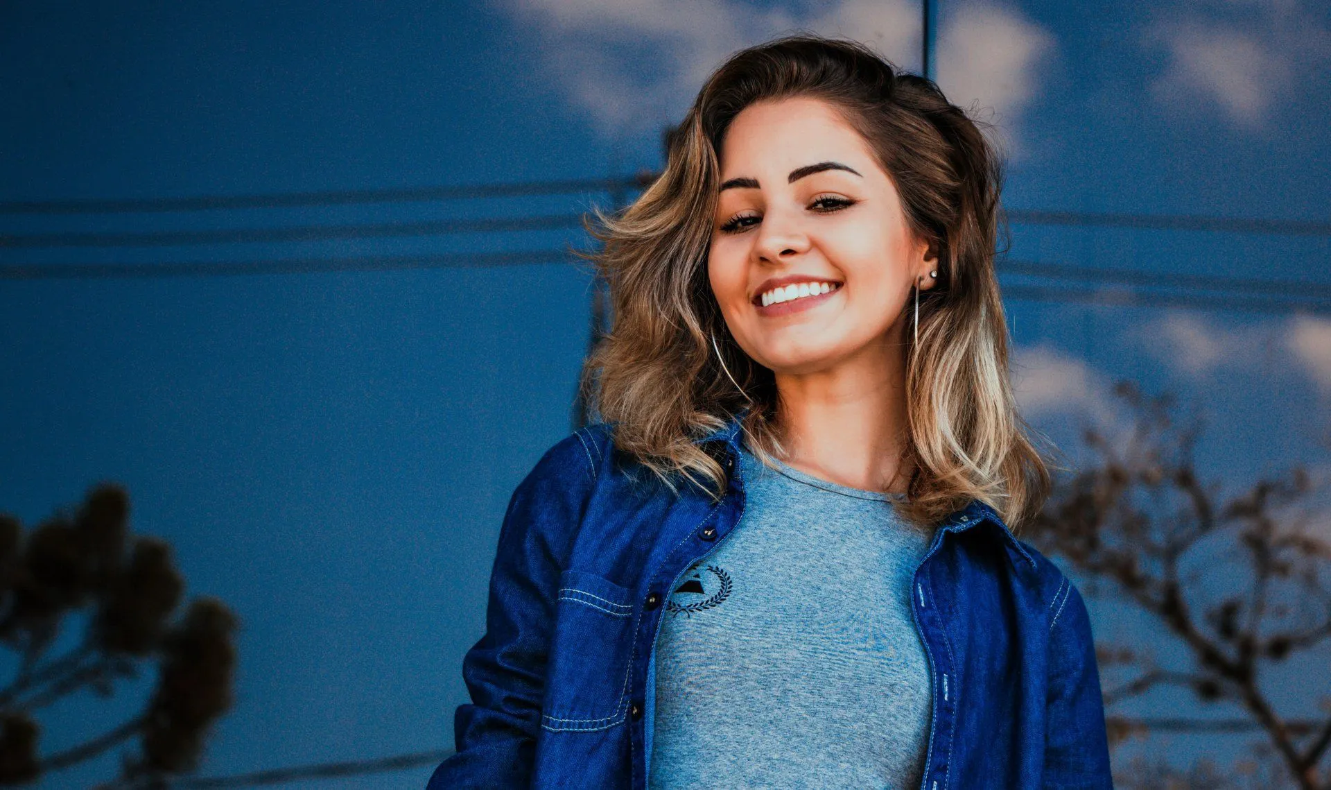 Smiling woman in a blue denim shirt stands in front of a reflective, blue surface.