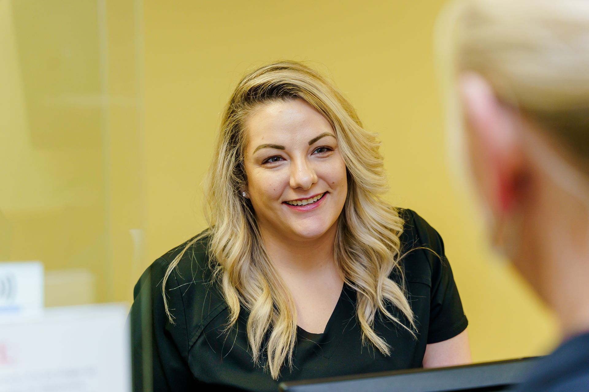 A woman with long blonde hair smiles while speaking to another person in an office setting.