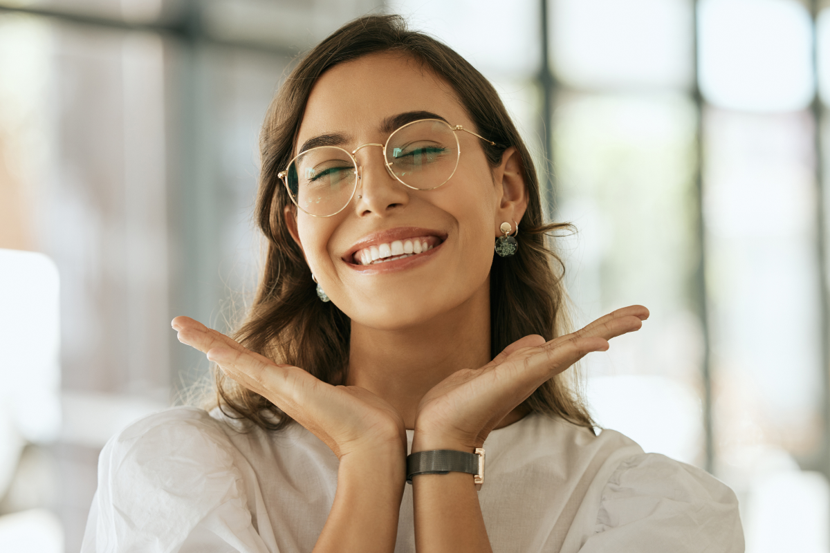 Smiling young woman with glasses and earrings posing with hands under her chin in a bright indoor setting.