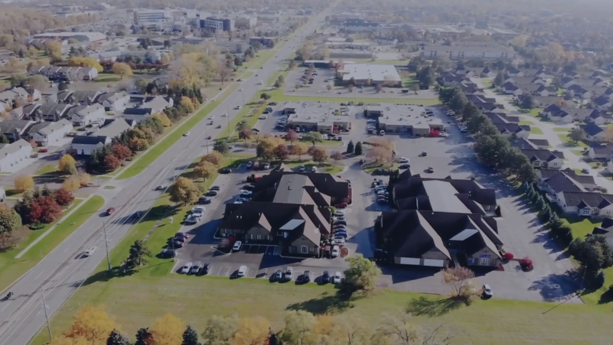 Aerial view of suburban area with residential houses, roads, parking lots, and buildings surrounded by trees with autumn foliage.