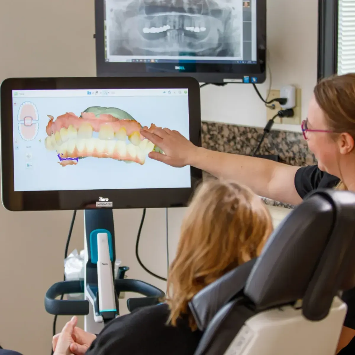Dental professional showing a digital 3D model of teeth on a screen to a patient in a dental chair.