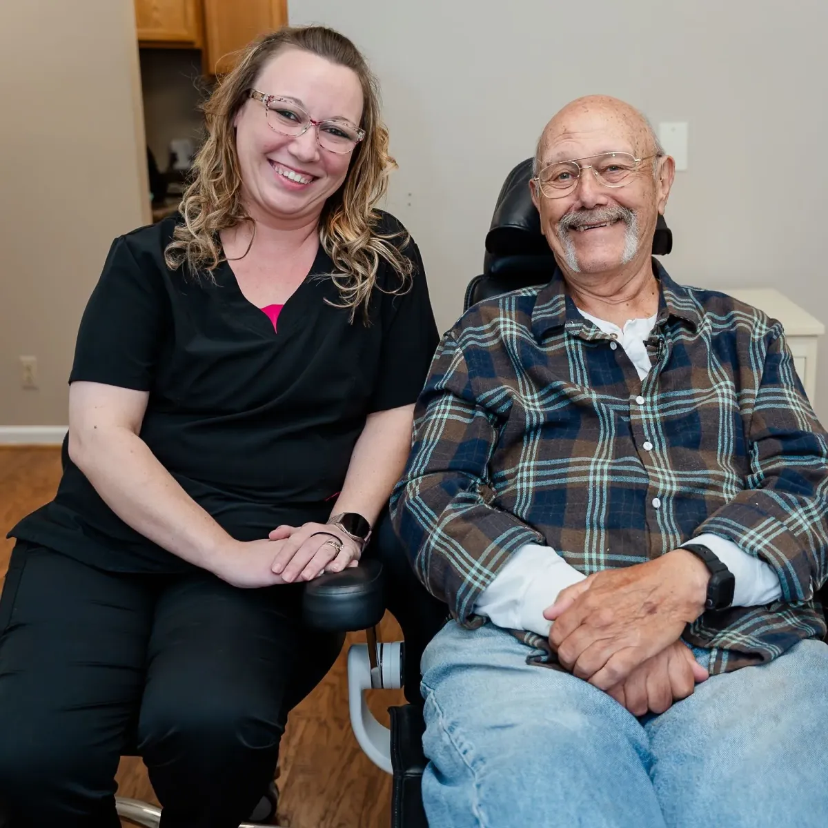 Smiling woman in black scrubs sitting next to a smiling elderly man in a wheelchair wearing a plaid shirt and jeans.