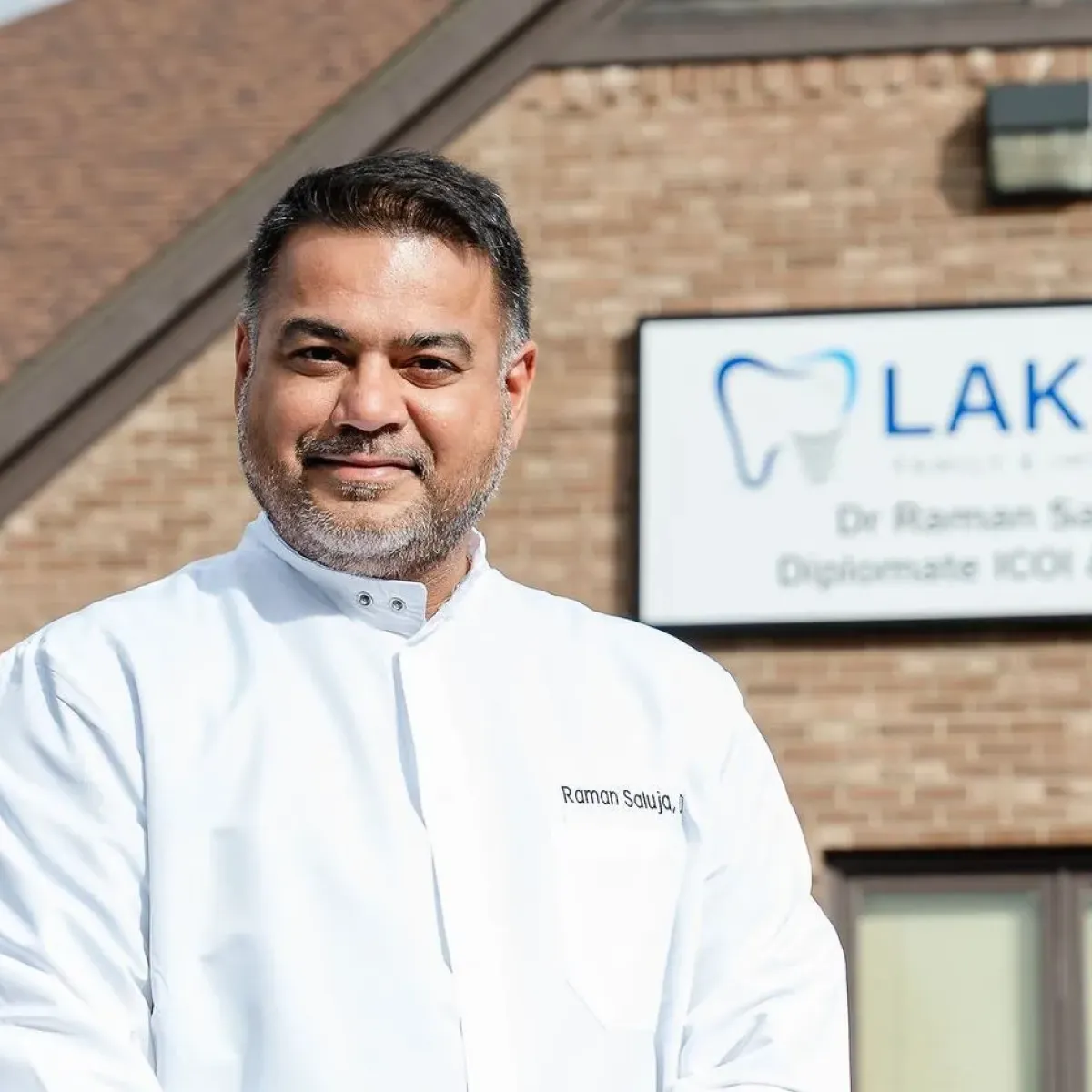 Smiling male dentist wearing a white coat standing outdoors in front of a dental clinic sign with a tooth logo.