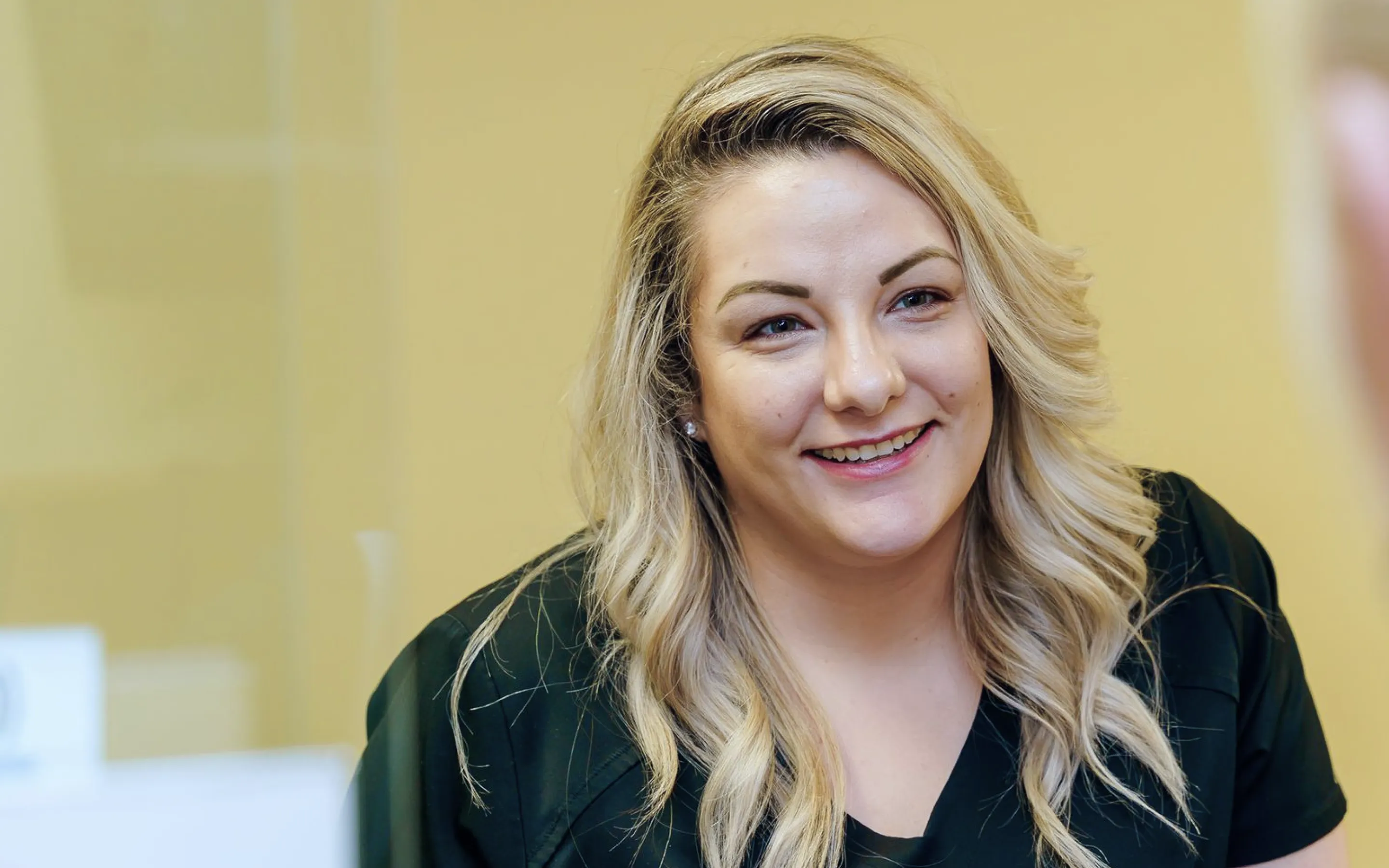 Smiling woman with blonde wavy hair wearing a black top against a plain yellow background.