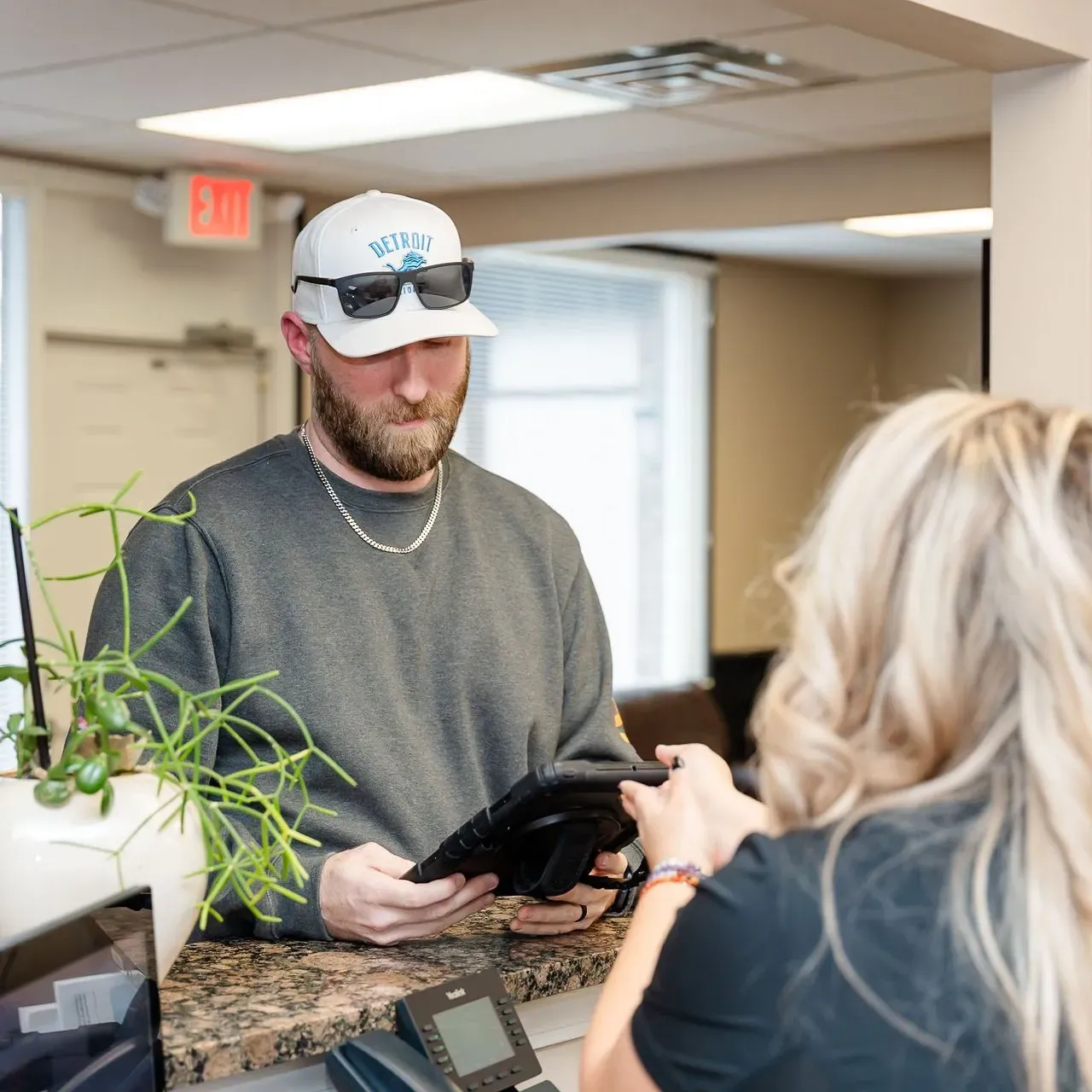 Man wearing a Detroit cap and sunglasses interacts with a woman handing him a device over a reception desk.