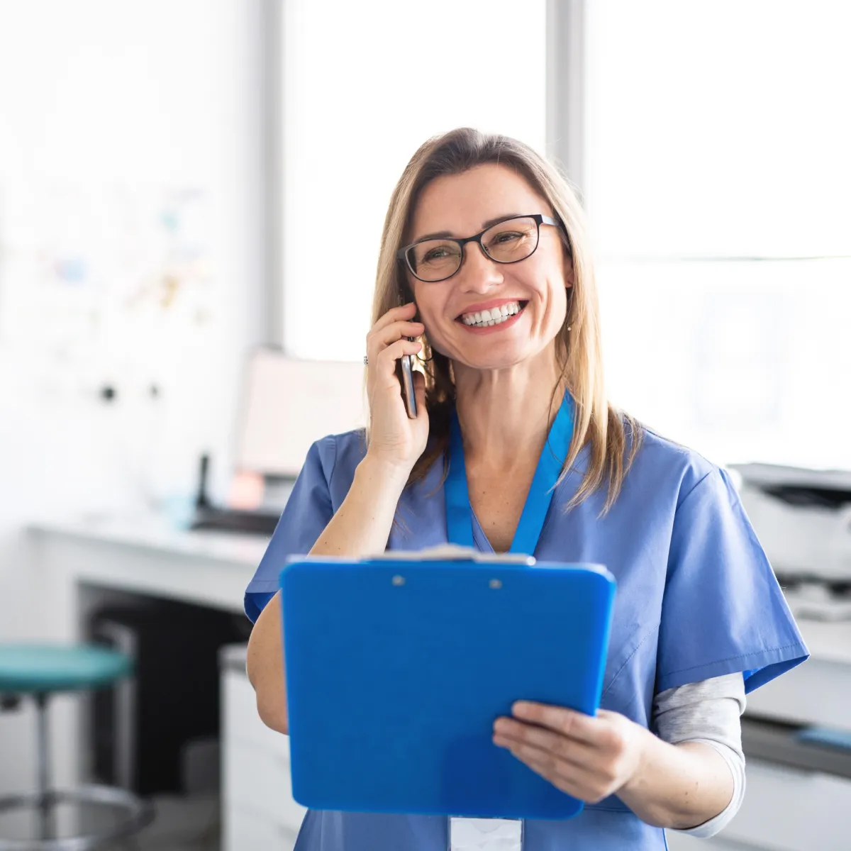 Smiling female healthcare professional in blue scrubs holding a clipboard and talking on a phone in a medical office.