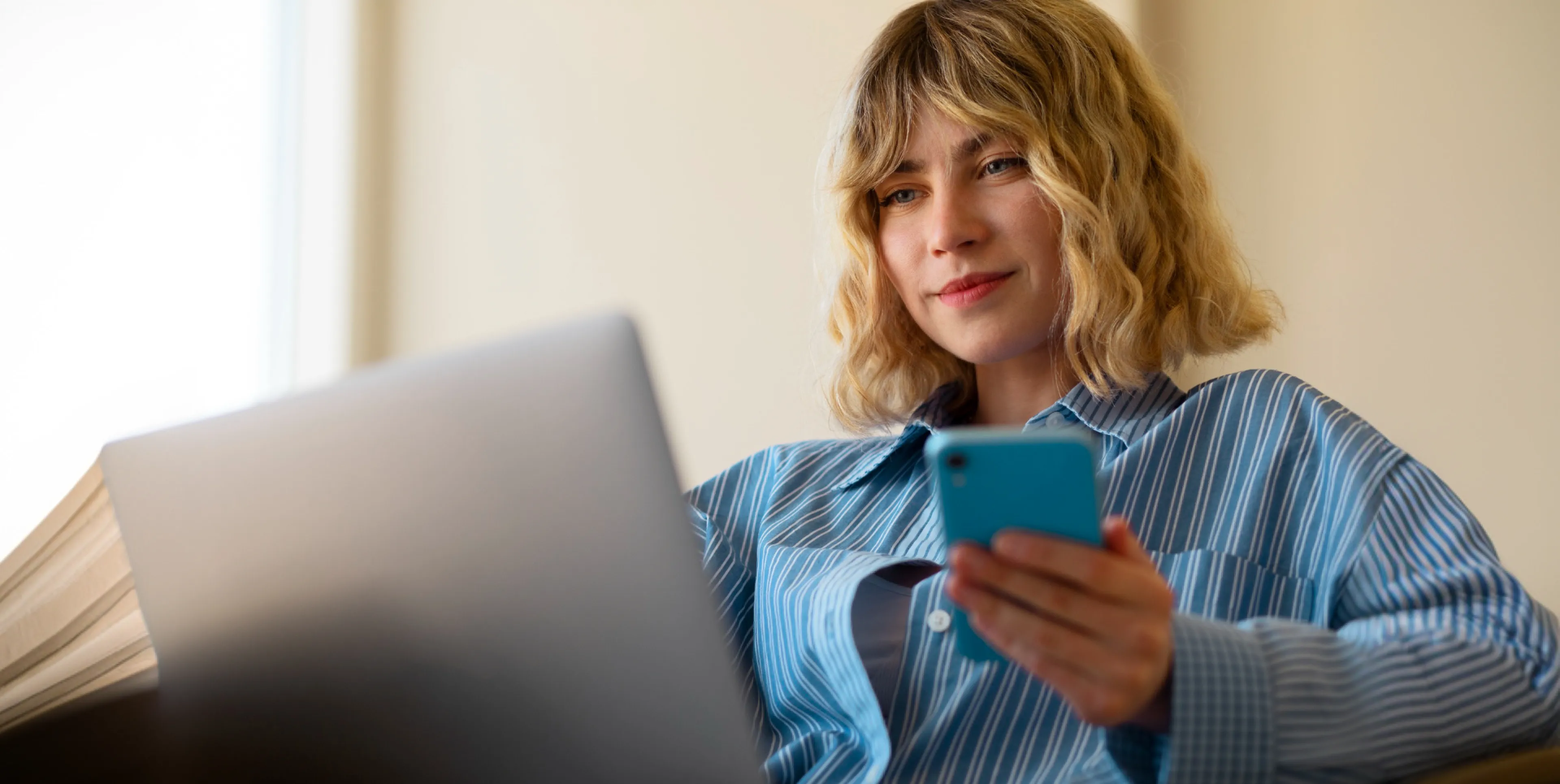 Young woman with blonde hair using a smartphone and a laptop, smiling gently.