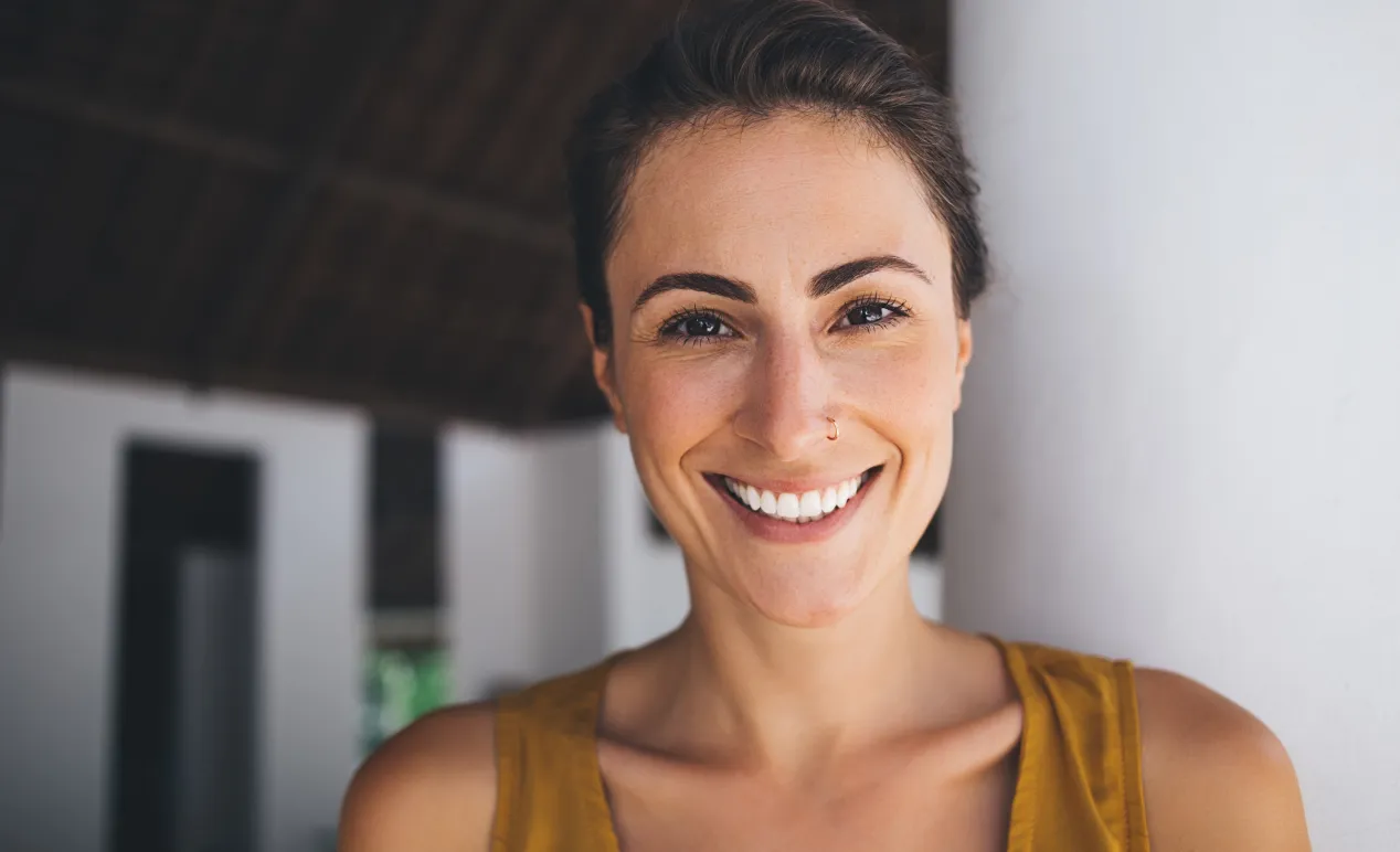 Smiling woman with a nose ring showing bright white teeth, wearing a mustard yellow top.
