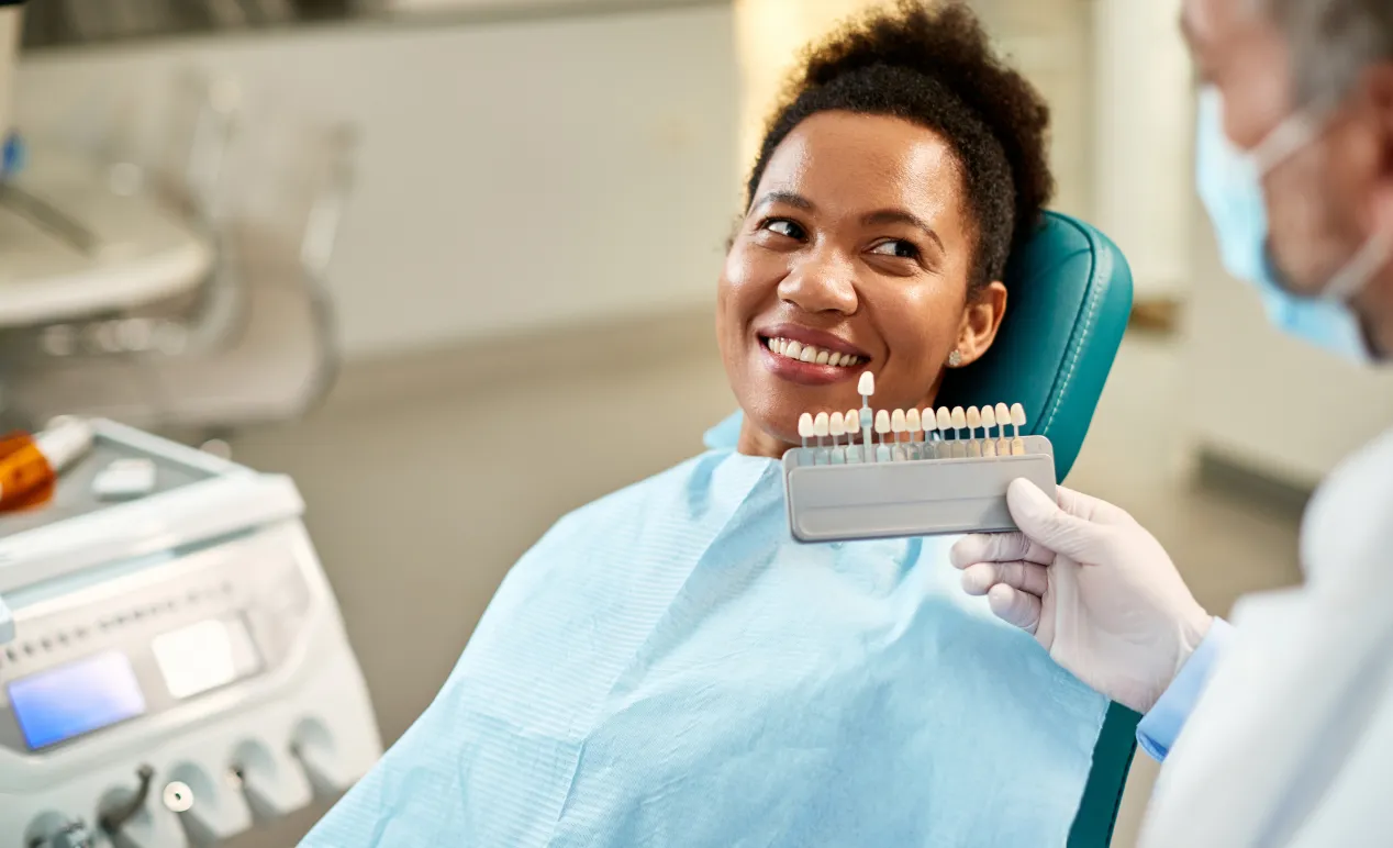 Smiling woman in dental chair comparing tooth color with a shade guide held by a dentist wearing gloves and a mask.