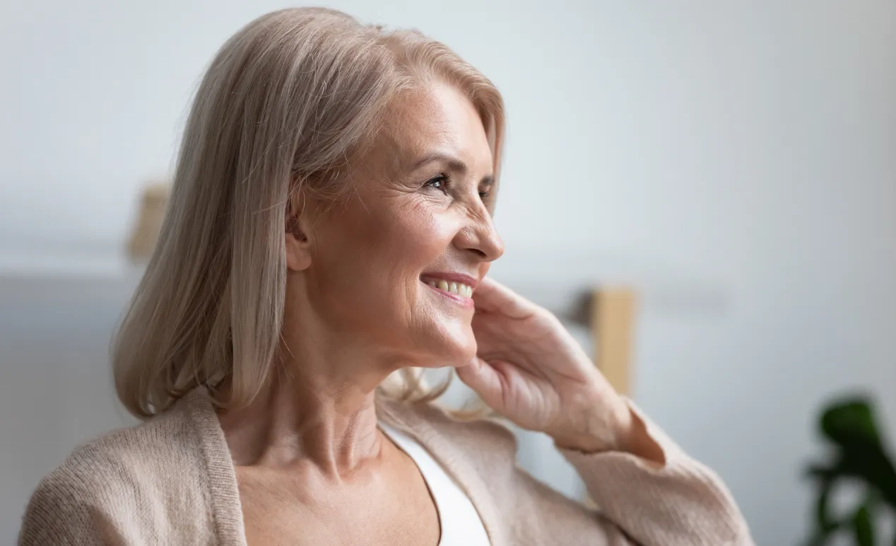 Smiling middle-aged woman with light blonde hair gazing to the side, resting her hand on her neck.