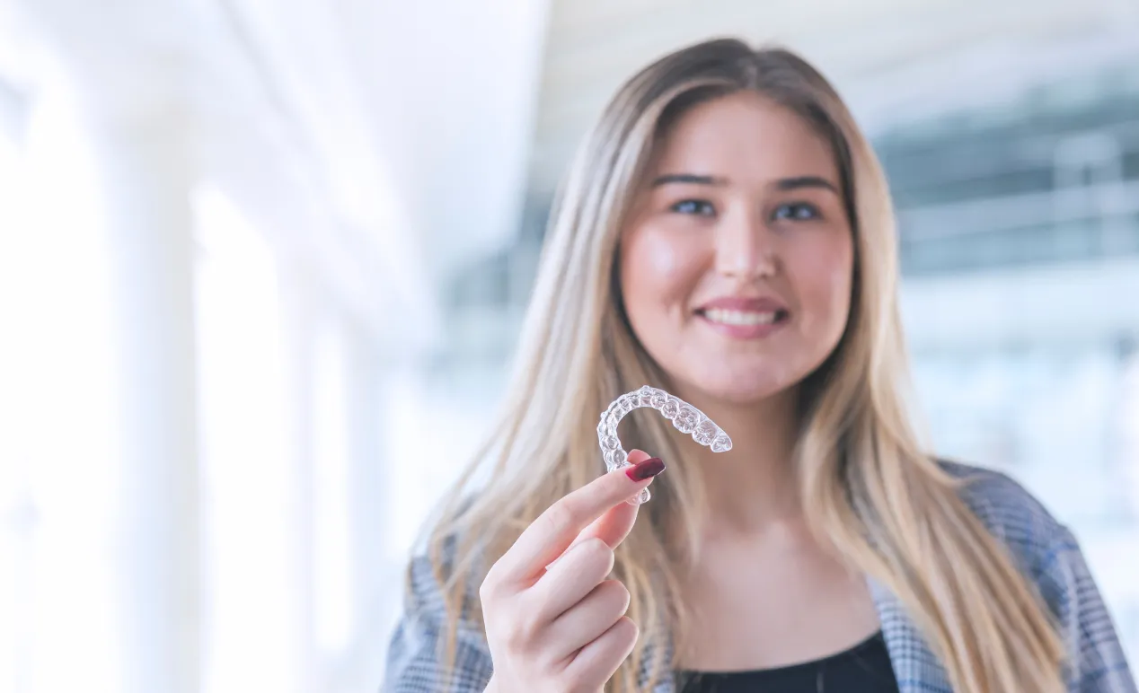 Smiling woman holding a clear dental aligner in her hand indoors.