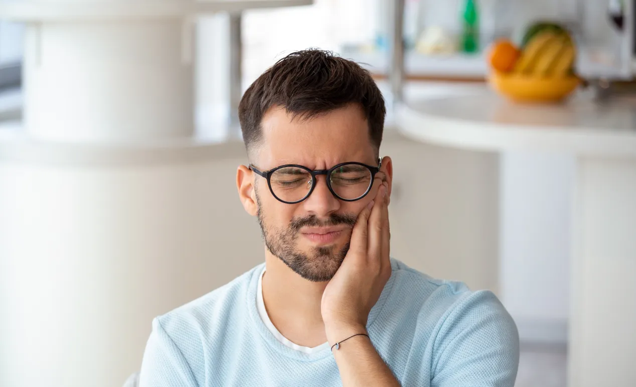 Man wearing glasses in light blue shirt holding his cheek with a pained expression indoors.