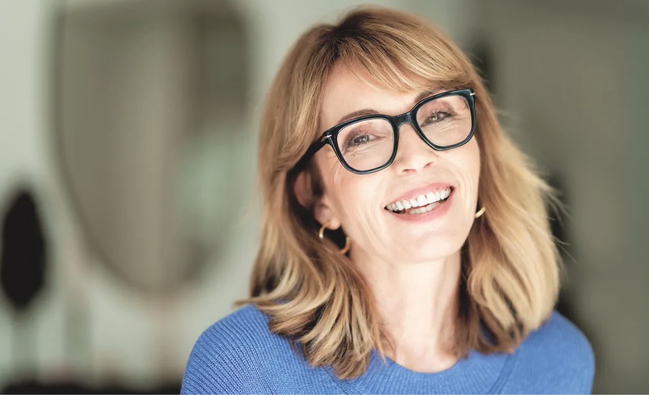 Smiling middle-aged woman with blonde hair, black glasses, and a blue sweater in a softly lit room.