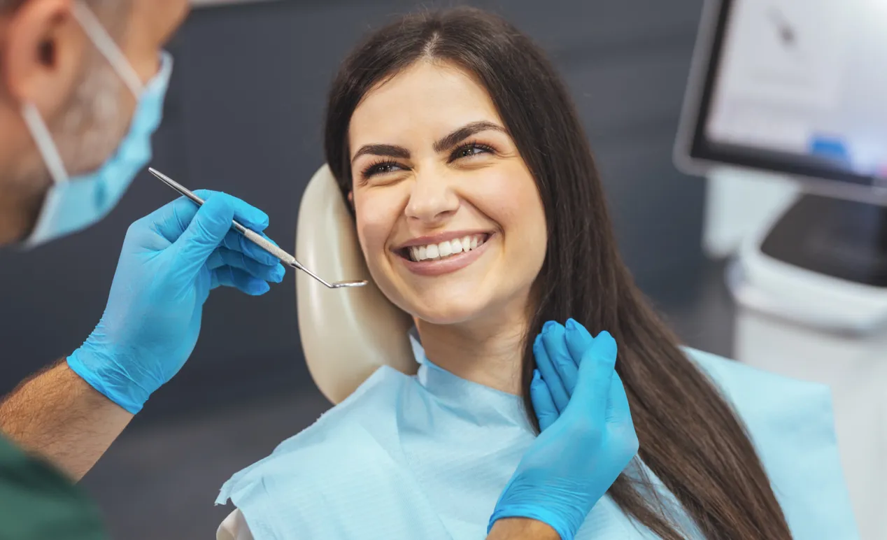 Smiling woman sitting in dental chair while dentist wearing blue gloves holds dental mirror near her mouth.
