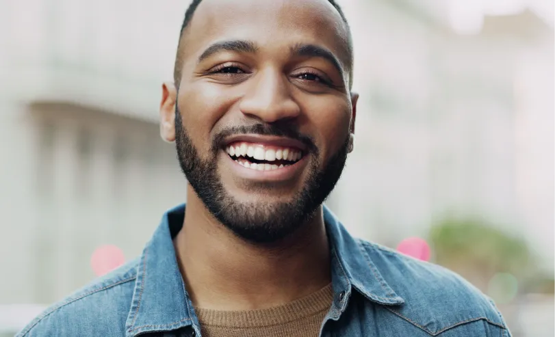 Smiling man with beard wearing a denim jacket and brown sweater outdoors in an urban setting.