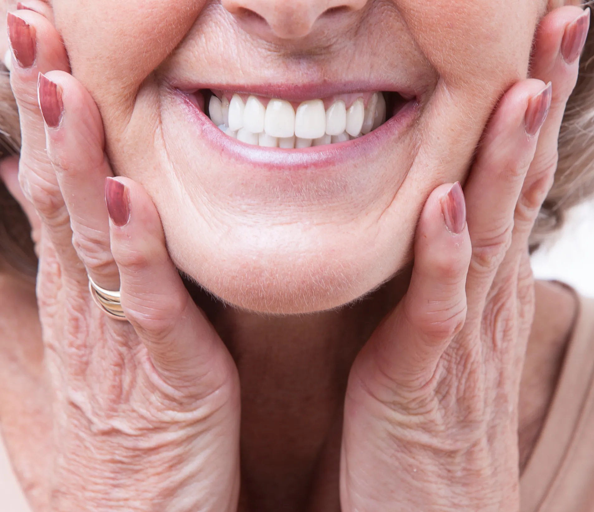Close-up of an elderly woman smiling, holding her face with both hands showing painted nails and visible wrinkles.