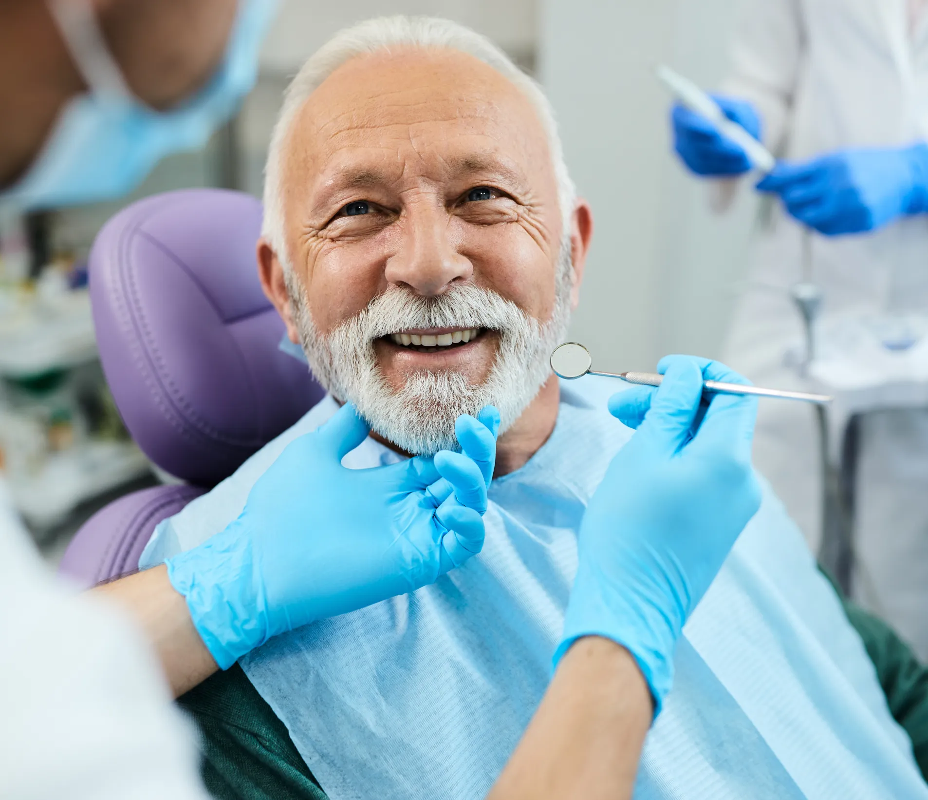 Smiling elderly man with white beard sitting in a dental chair while a dentist wearing blue gloves examines his teeth with dental tools.