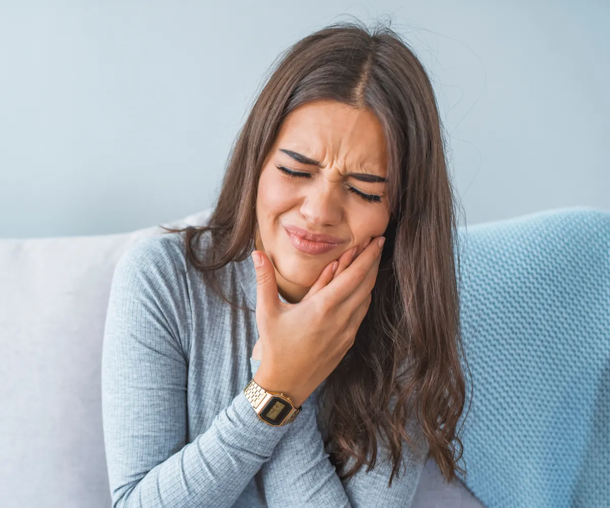 Young woman sitting on a couch with eyes closed, holding her jaw in pain, showing signs of a toothache.
