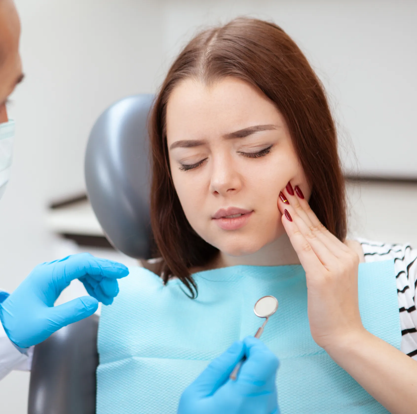 Young woman in dental chair holding her cheek in pain while dentist wearing blue gloves holds a dental mirror.