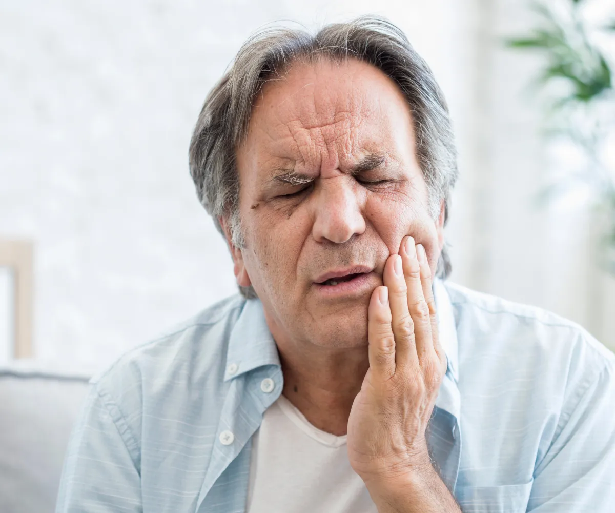 Middle-aged man with closed eyes holding his cheek in pain, indicating a toothache or facial discomfort.