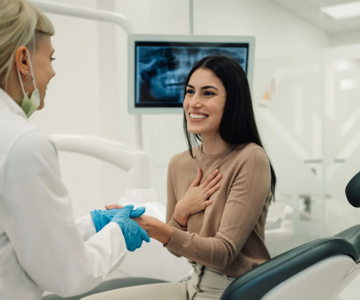 Smiling woman sitting in a dental chair holding hands with a gloved dentist in a clinic with a dental X-ray on a screen behind them.