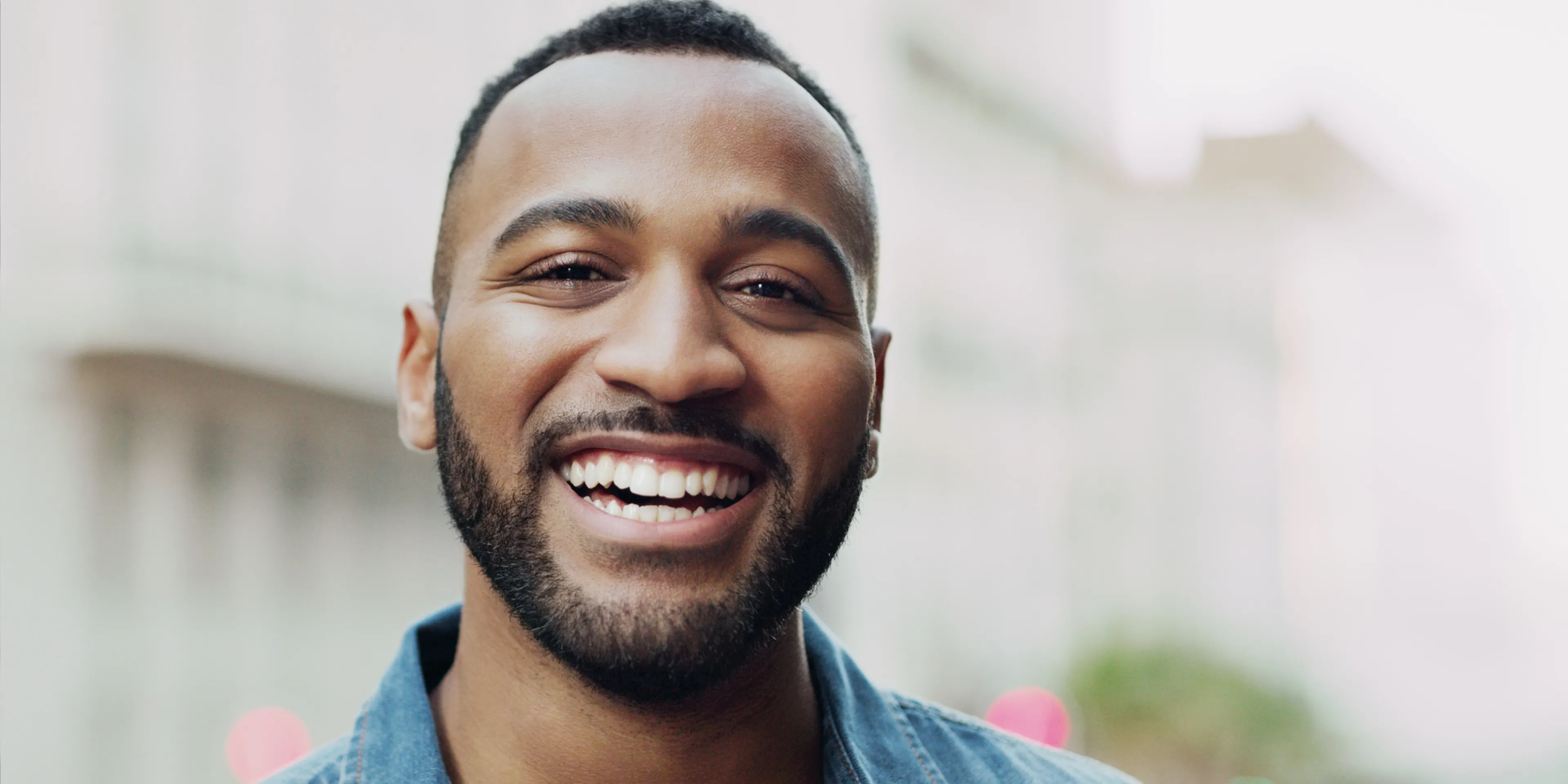 Smiling man with a beard showing white teeth outdoors in a blurred urban background.