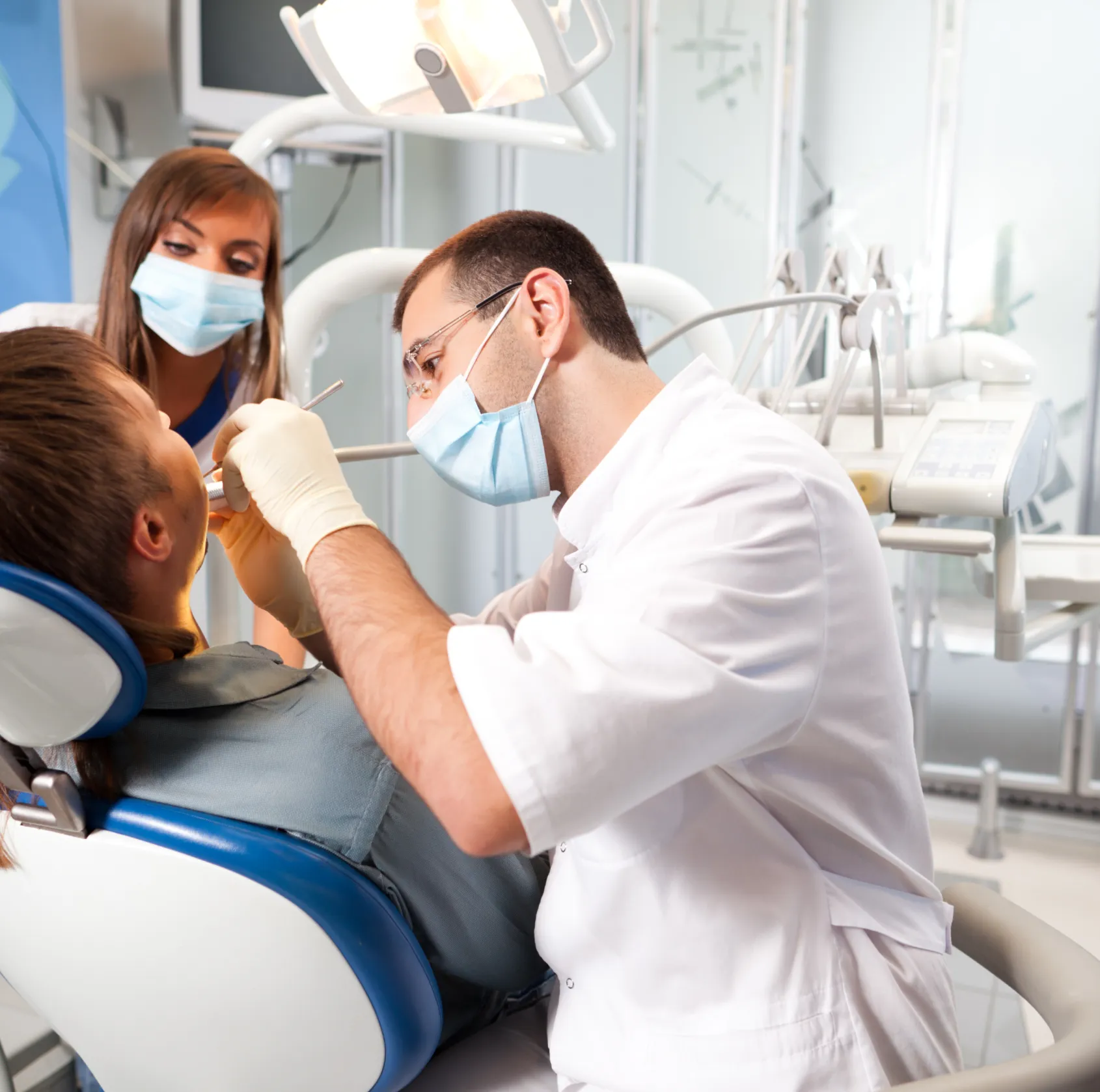 Dentist in white coat and mask examining a patient's teeth in a dental clinic with an assistant observing.