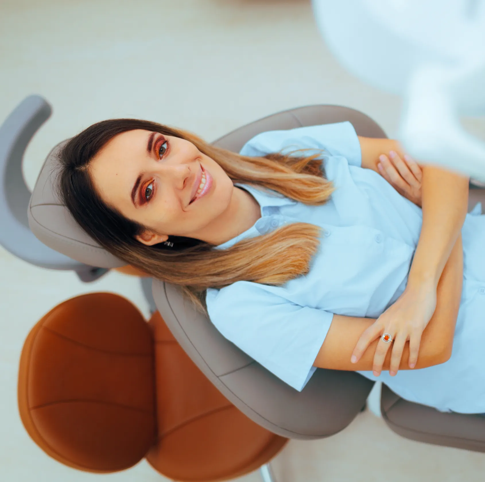 Smiling woman reclining in a dental chair with arms crossed, wearing a light blue shirt.