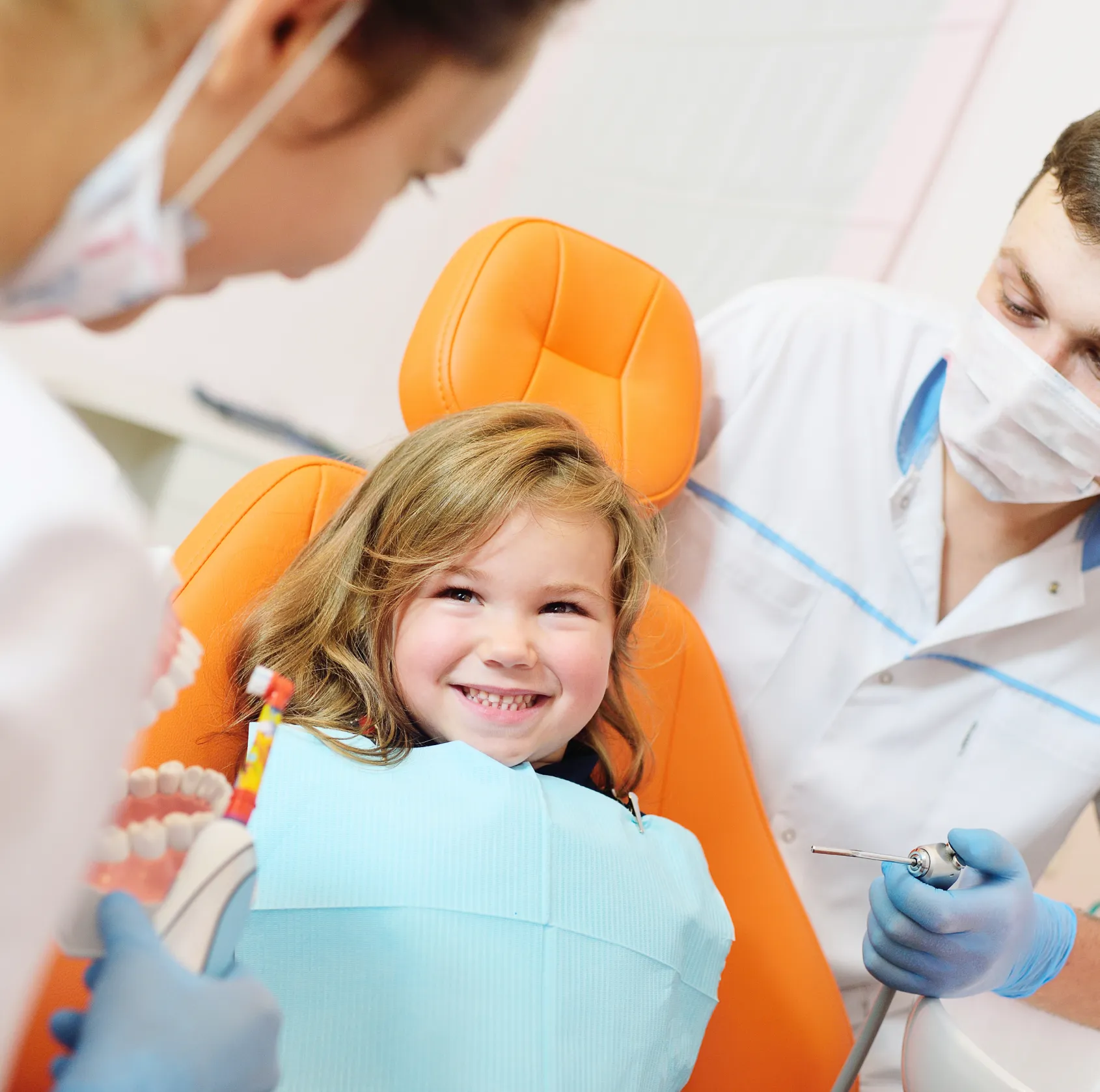 Smiling young girl sitting in orange dental chair during a dental checkup with two dentists wearing gloves and masks.