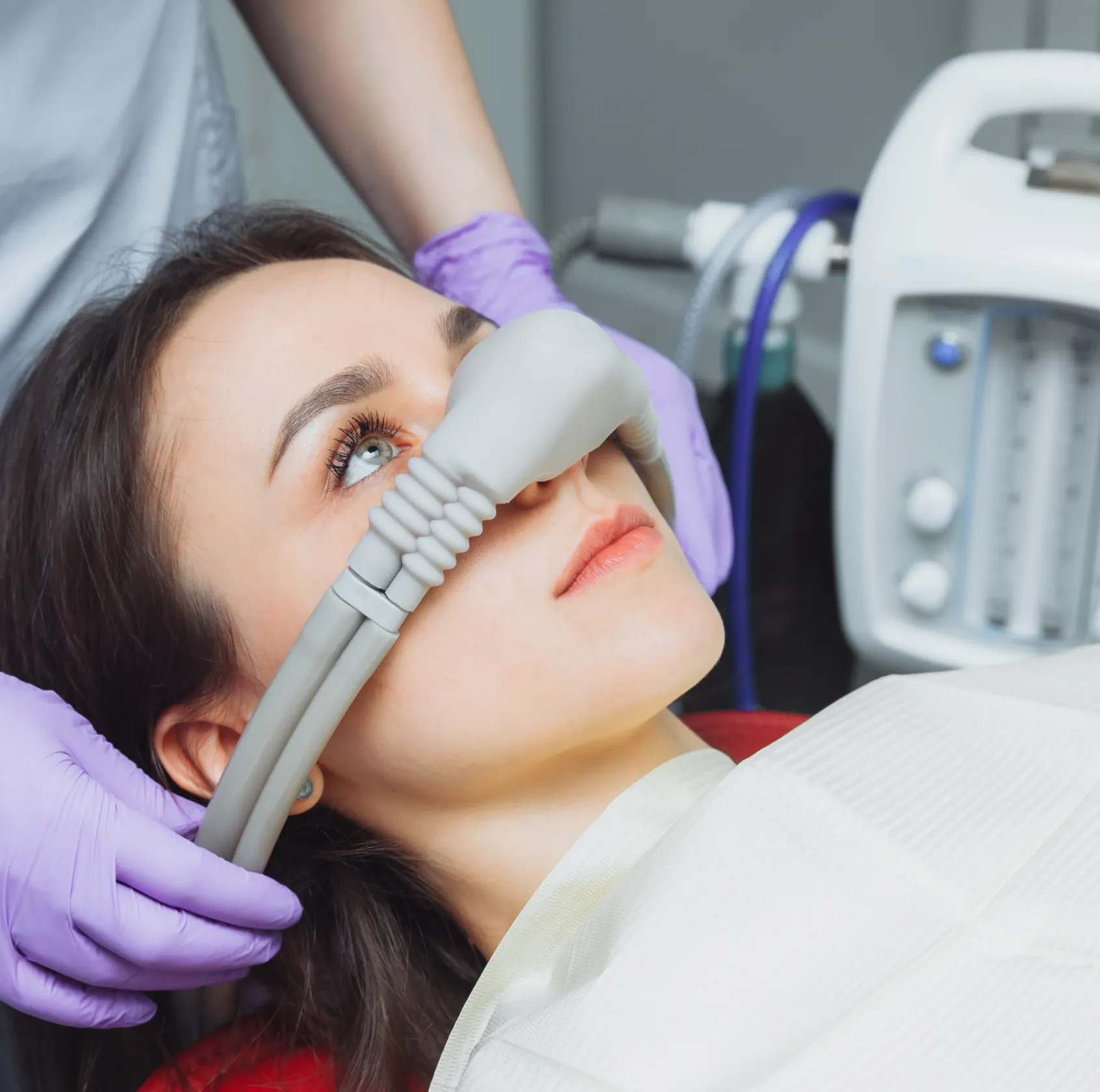 Woman lying down with a sedation mask over her nose, held by a medical professional wearing purple gloves.
