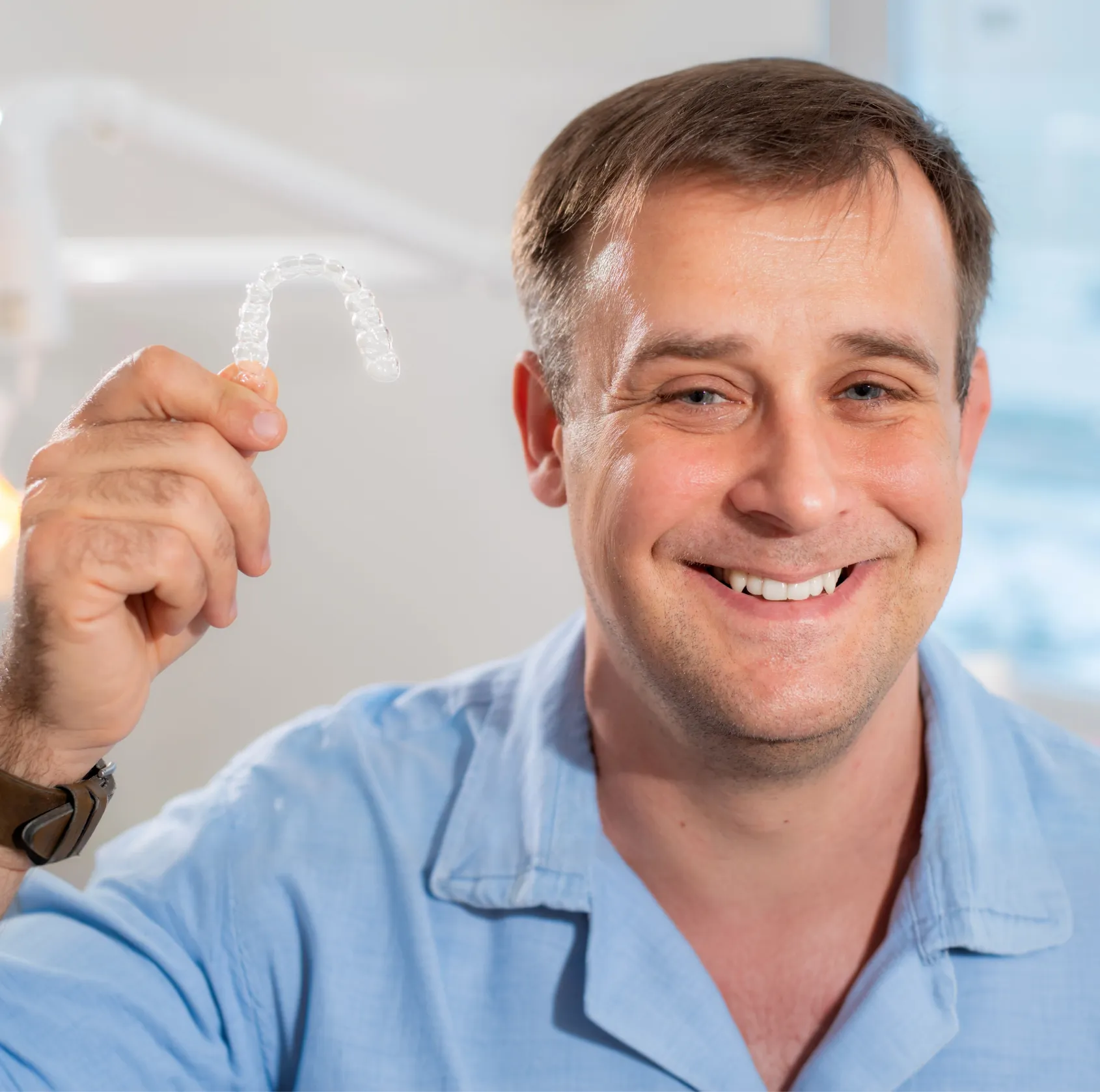 Smiling man in a light blue shirt holding a clear dental aligner in a dental office.