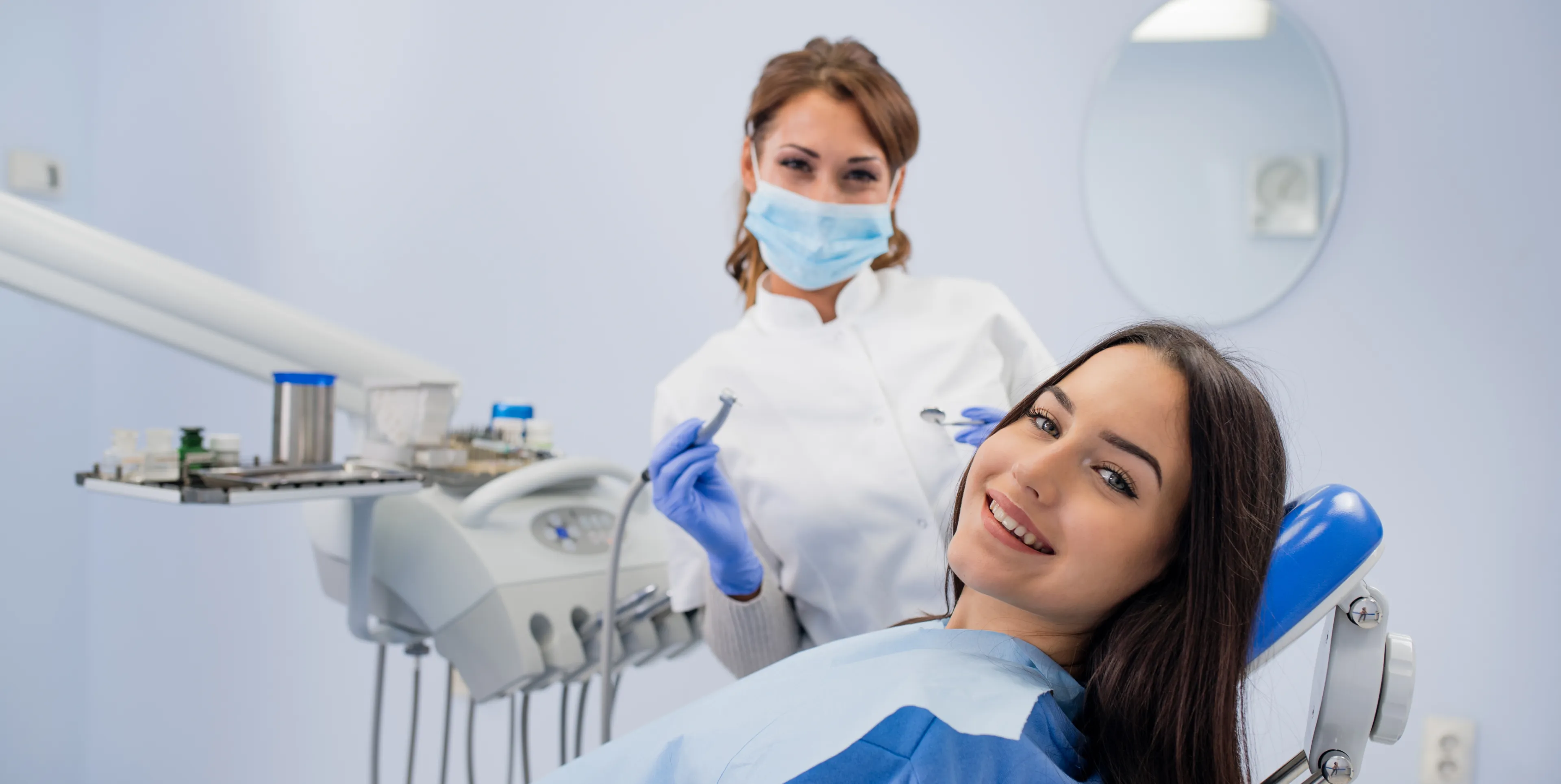 Smiling woman sitting in dental chair with a masked dentist holding dental tools in the background.