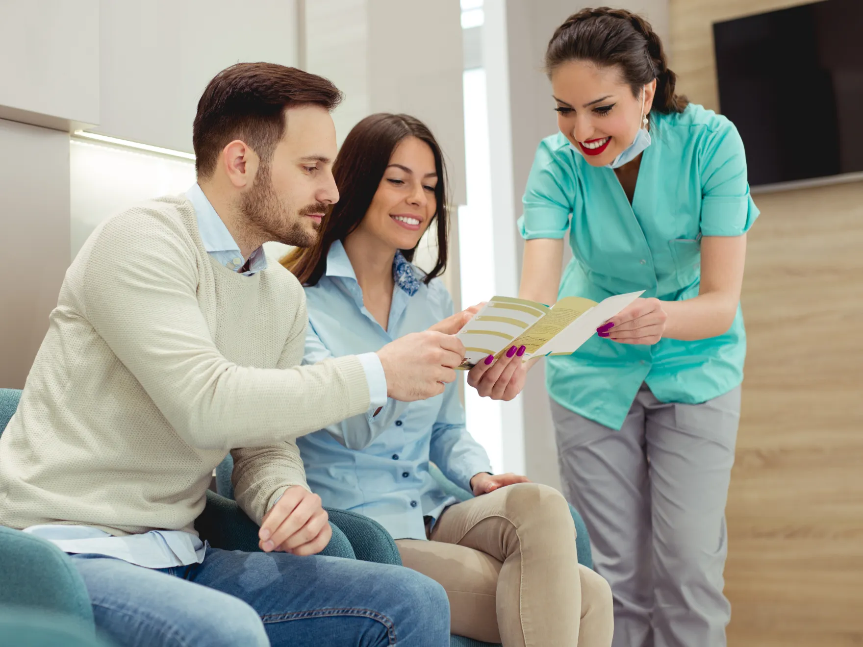 Healthcare professional in scrubs showing a brochure to a smiling couple seated in a clinic waiting area.