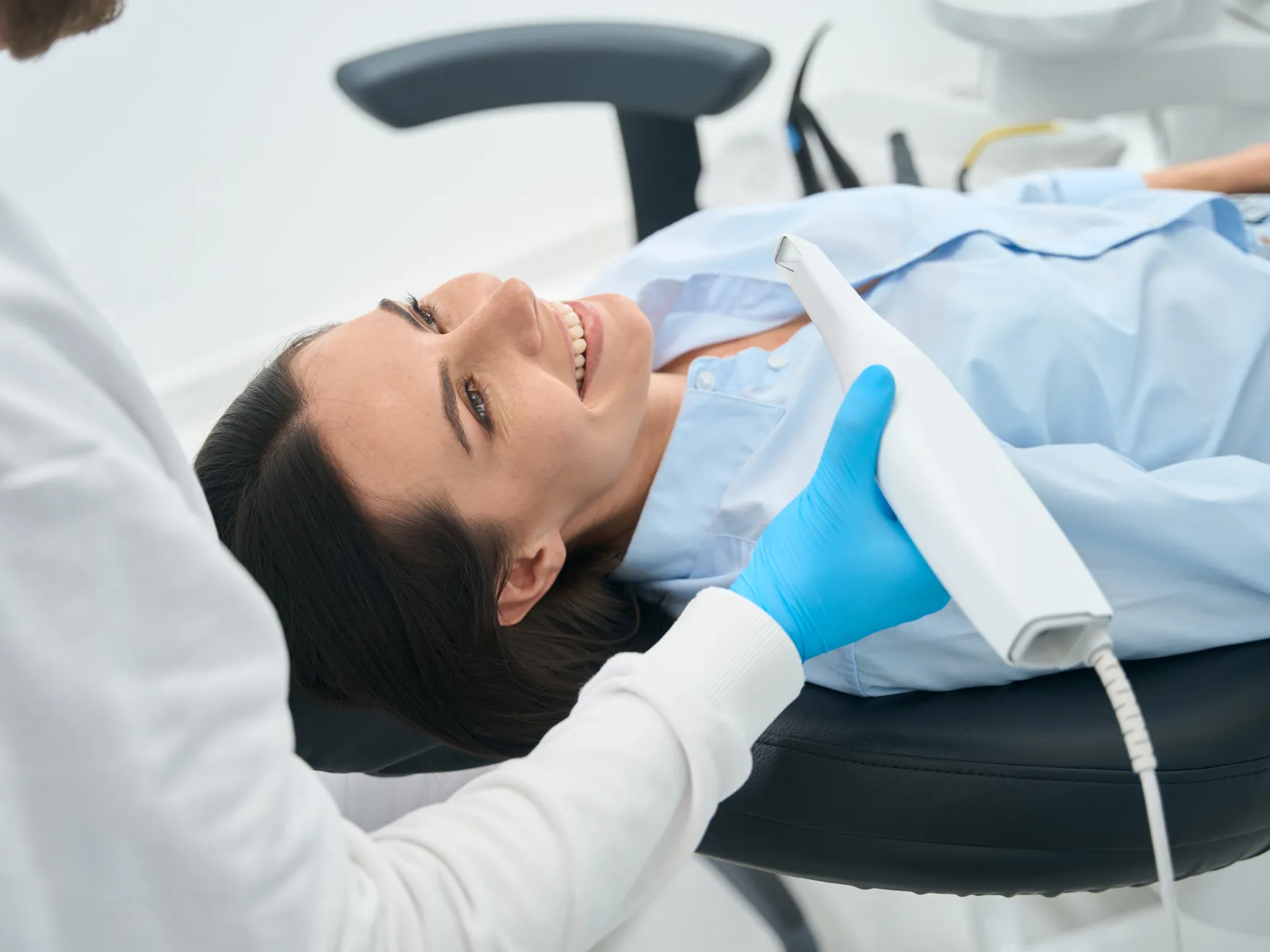 Smiling woman lying back in a dental chair while a dentist wearing blue gloves holds a dental scanner near her mouth.