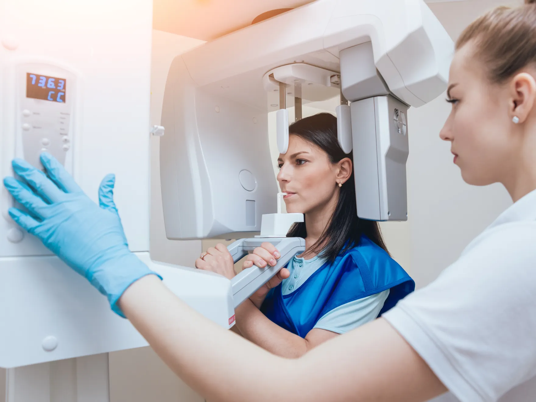 Woman wearing a protective lead apron undergoing a dental X-ray while technician adjusts the X-ray machine controls.