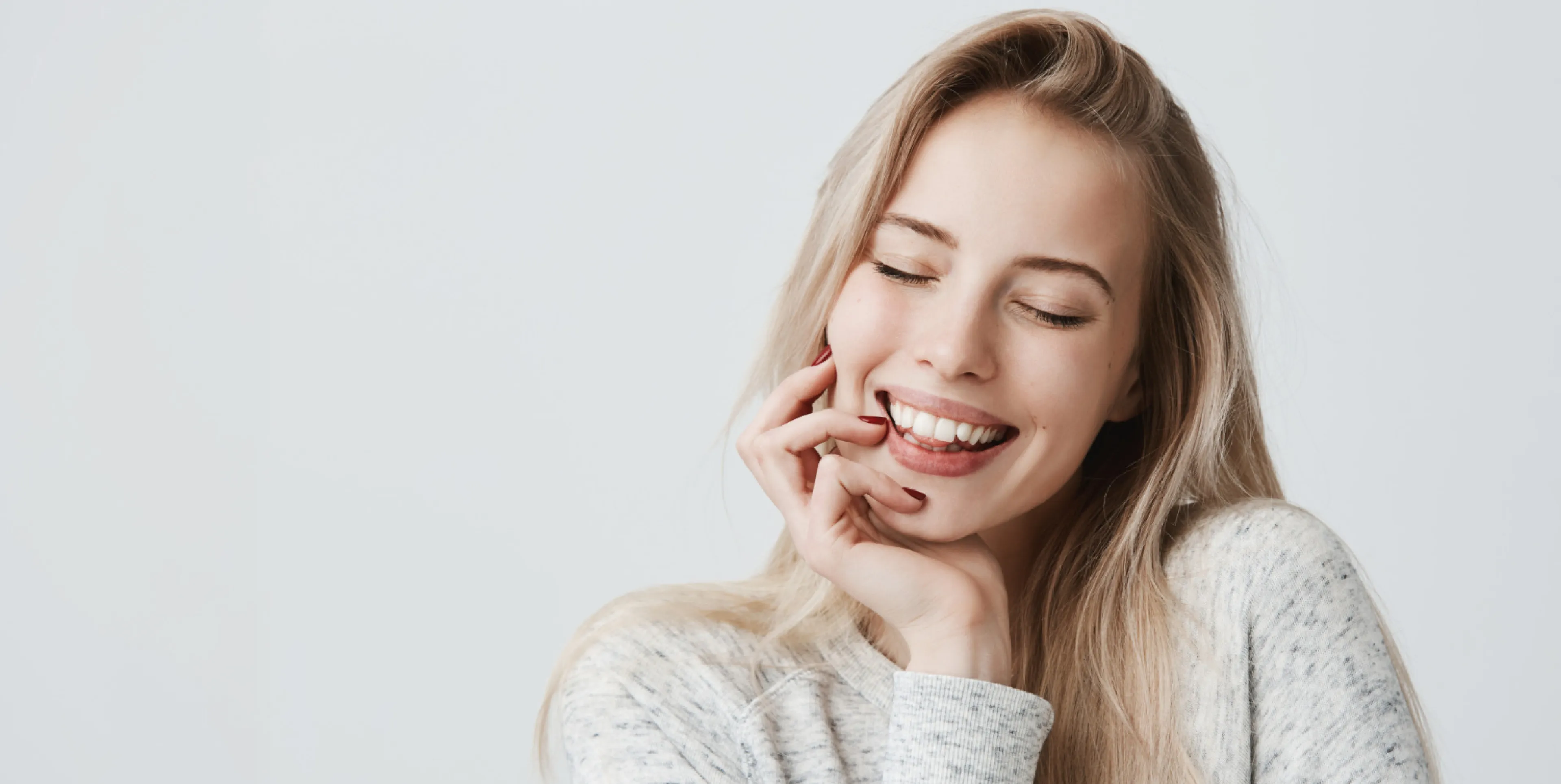 Close-up of a smiling young woman with long blonde hair, eyes closed, touching her face gently.