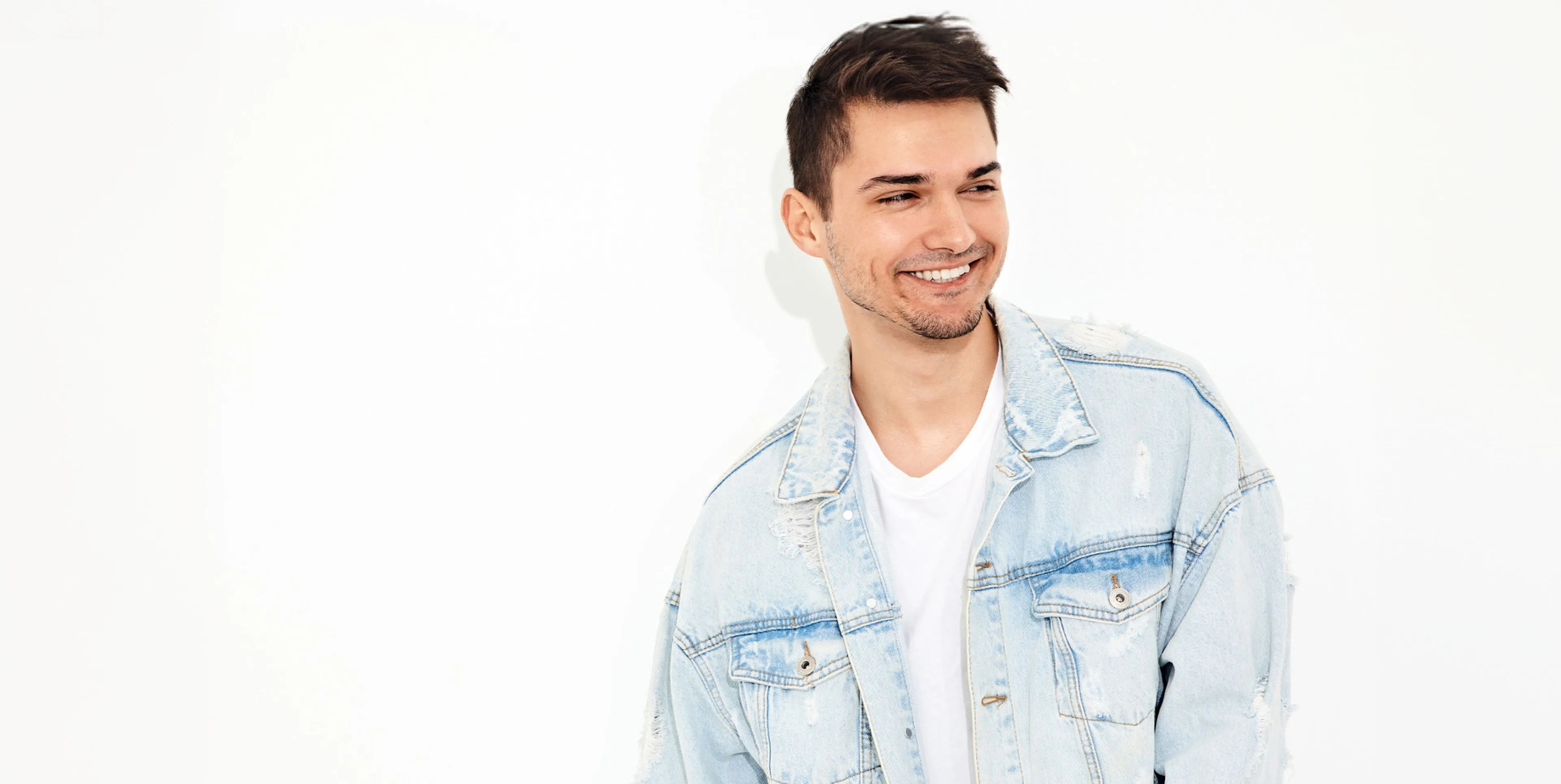 Smiling young man wearing a light blue denim jacket and a white t-shirt looking to the side against a plain white background.