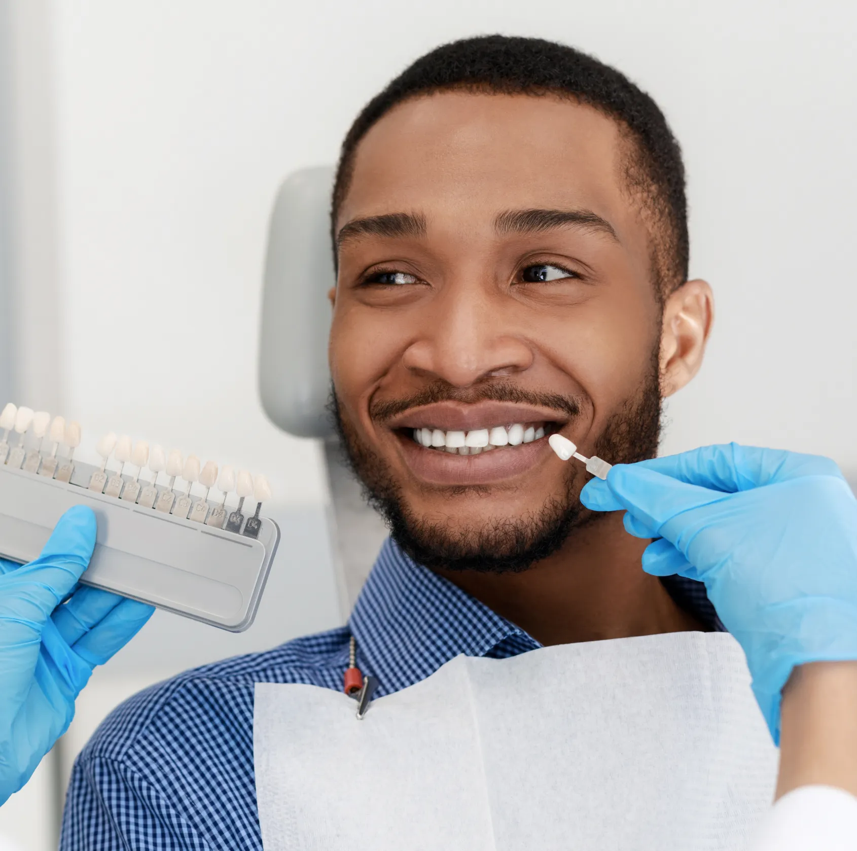 Dentist wearing blue gloves holding a dental shade guide next to a smiling man's teeth for color matching.