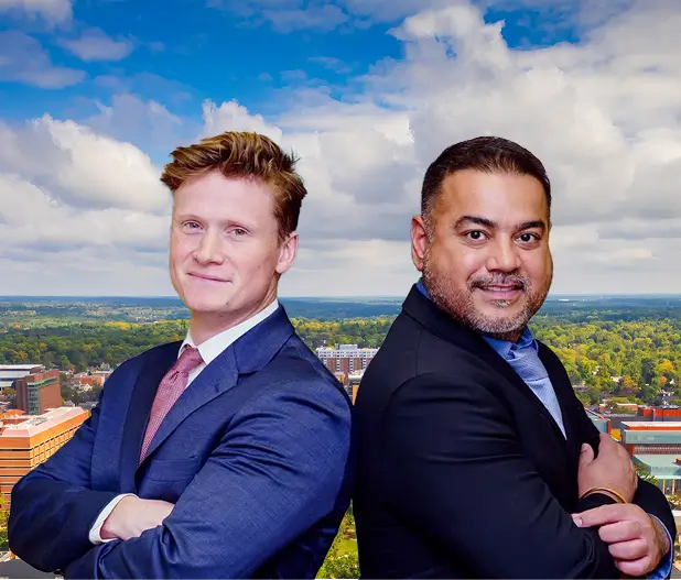 Two professionally dressed men standing back-to-back with arms crossed, smiling against a cityscape and partly cloudy sky background.