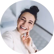 Smiling young woman with hair in a bun wearing a white top, taking a selfie indoors.