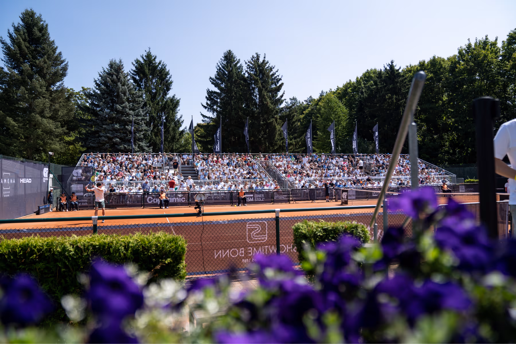 Tennisspieler, der auf einem Sandplatz schlägt, vor einer voll besetzten Tribüne mit Zuschauern und Bäumen im Hintergrund.