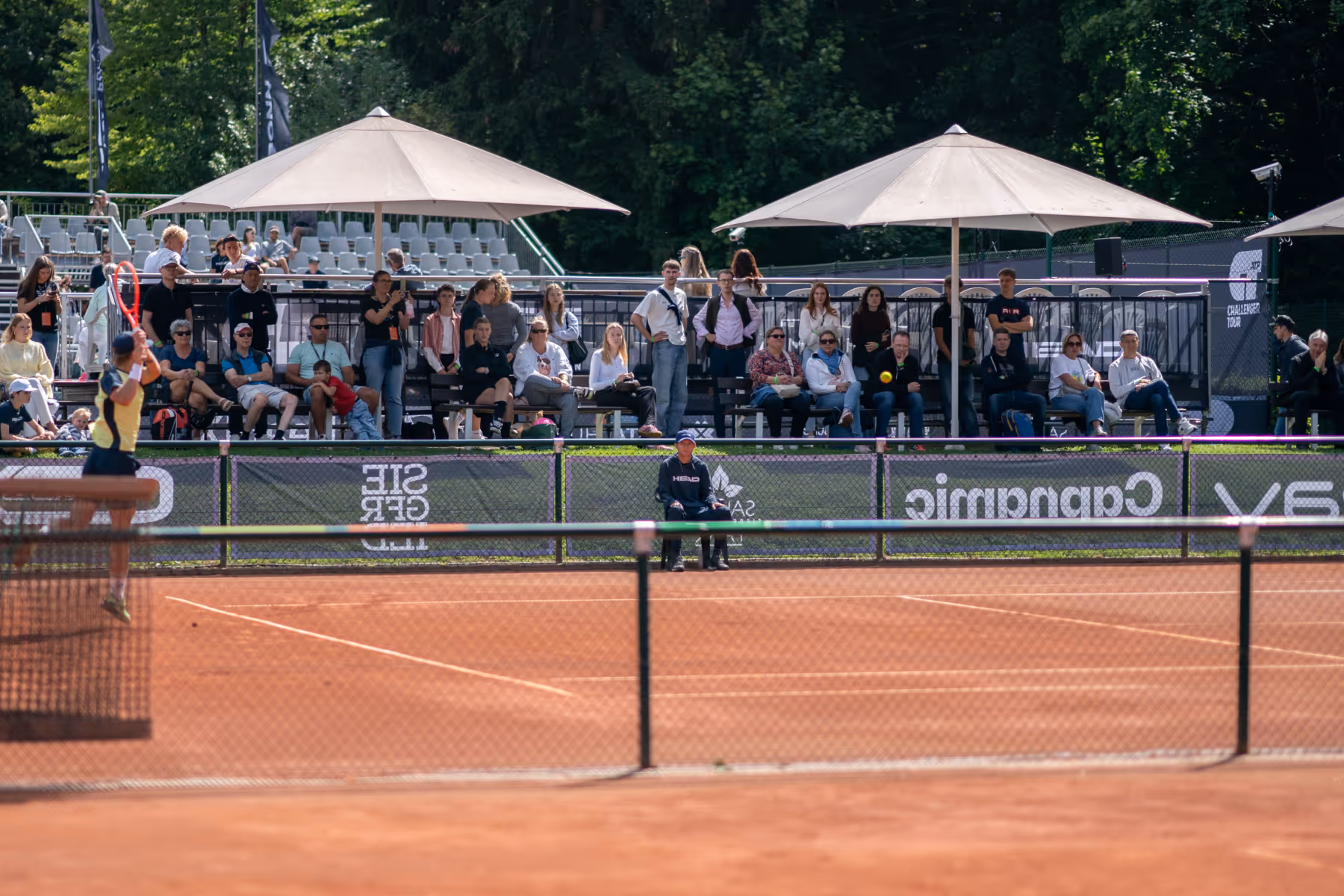 Tennisplatz mit Spielern im Match, Zuschauer auf Tribüne unter Sonnenschirmen und ein Schiedsrichter auf einem Stuhl.
