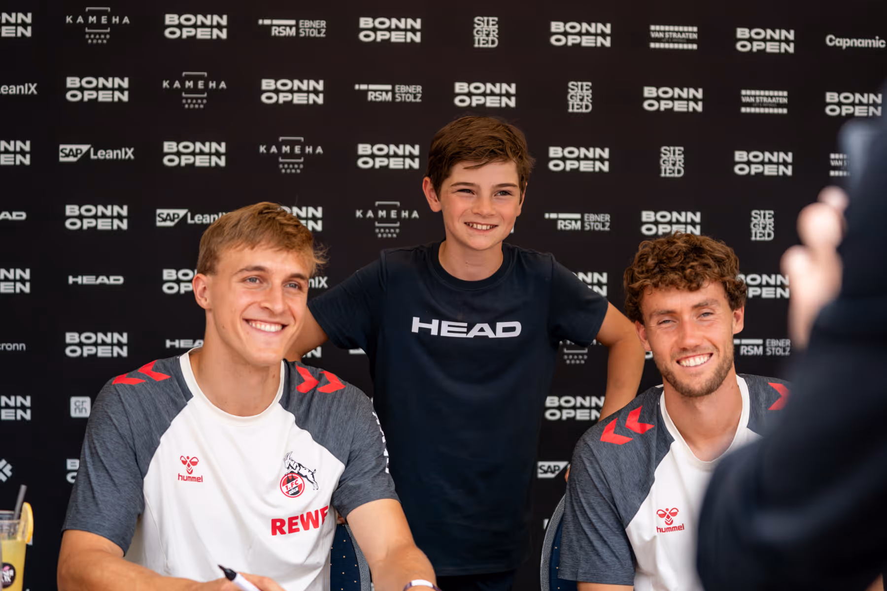 Zwei junge Männer in Sporttrikots sitzen und lächeln, während ein Kind mit einem HEAD-T-Shirt zwischen ihnen steht vor einer Bonn Open Sponsorenwand.